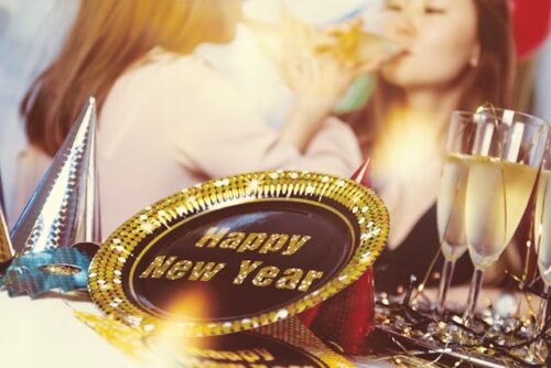 A festive scene unfolds with two people celebrating New Year's Eve. In the foreground, a decorative plate displays "Happy New Year" amidst party hats and champagne glasses. The background captures the joyous moment of someone sipping from a flute glass.