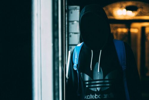 A person wearing a hoodie with a large logo on the front stands in dim lighting, their face obscured by shadows. With a blue backpack slung over one shoulder, they linger near a door with illuminated panels, casting an enigmatic aura possibly tied to a shoplifting charge.