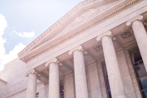 Low-angle view of a neoclassical building with tall, white marble columns reminiscent of ancient justice halls. The roof features an ornate pediment, and the sky is clear with a few clouds, bathed in sunlight.