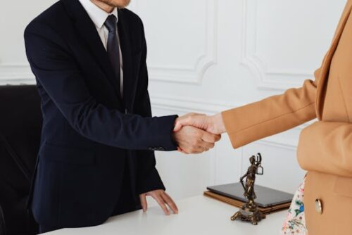 A man in a dark suit, likely a criminal defense attorney, shakes hands with a woman in a brown coat across the desk. A small statue and some folders are visible on the desk. The setting appears to be a professional office with white walls.